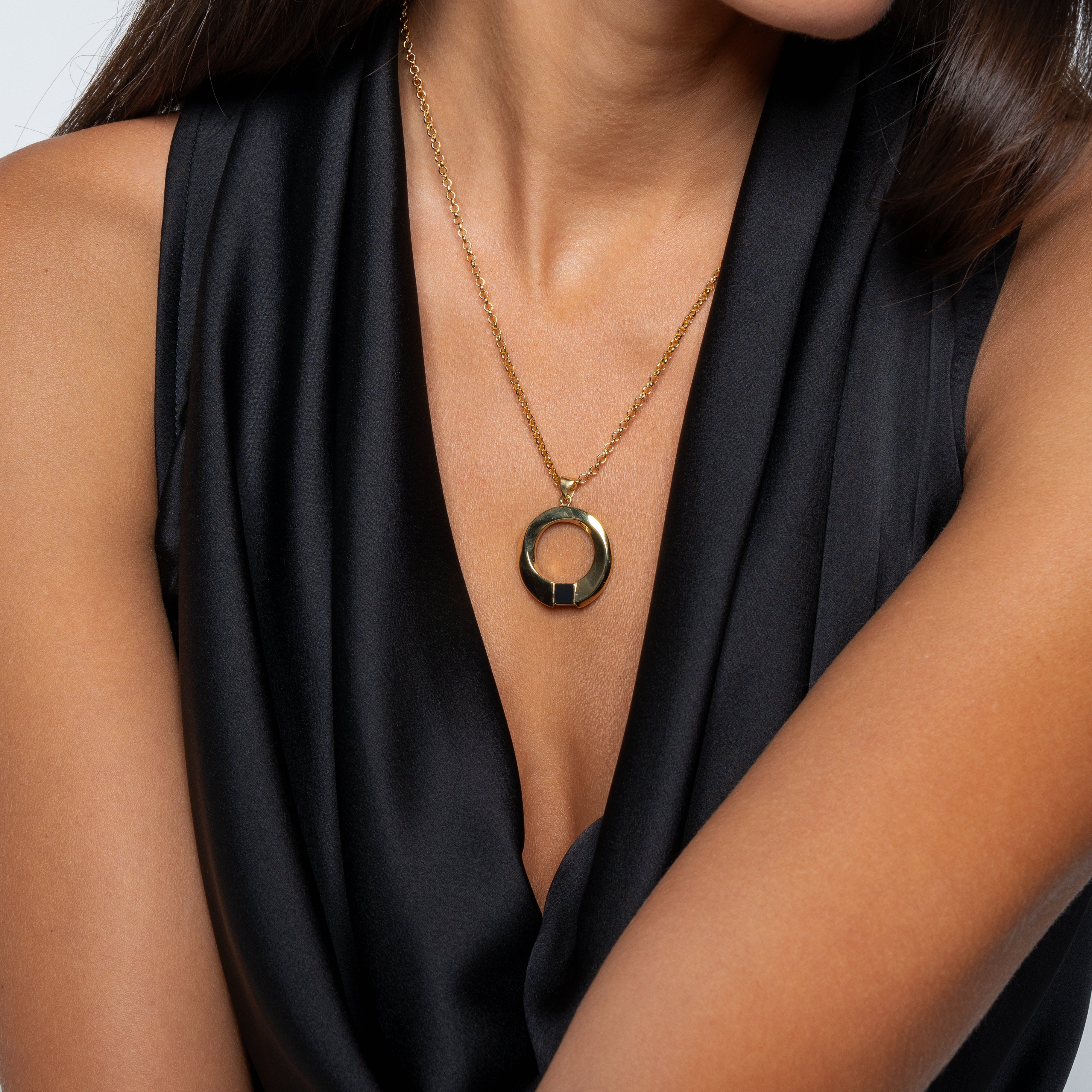 A woman in a sleeveless black satin top shows off the gold Eternity Circle Necklace from NanoBless Christian Jewelry, featuring a large circular pendant; her arms are crossed and part of her face is out of frame.