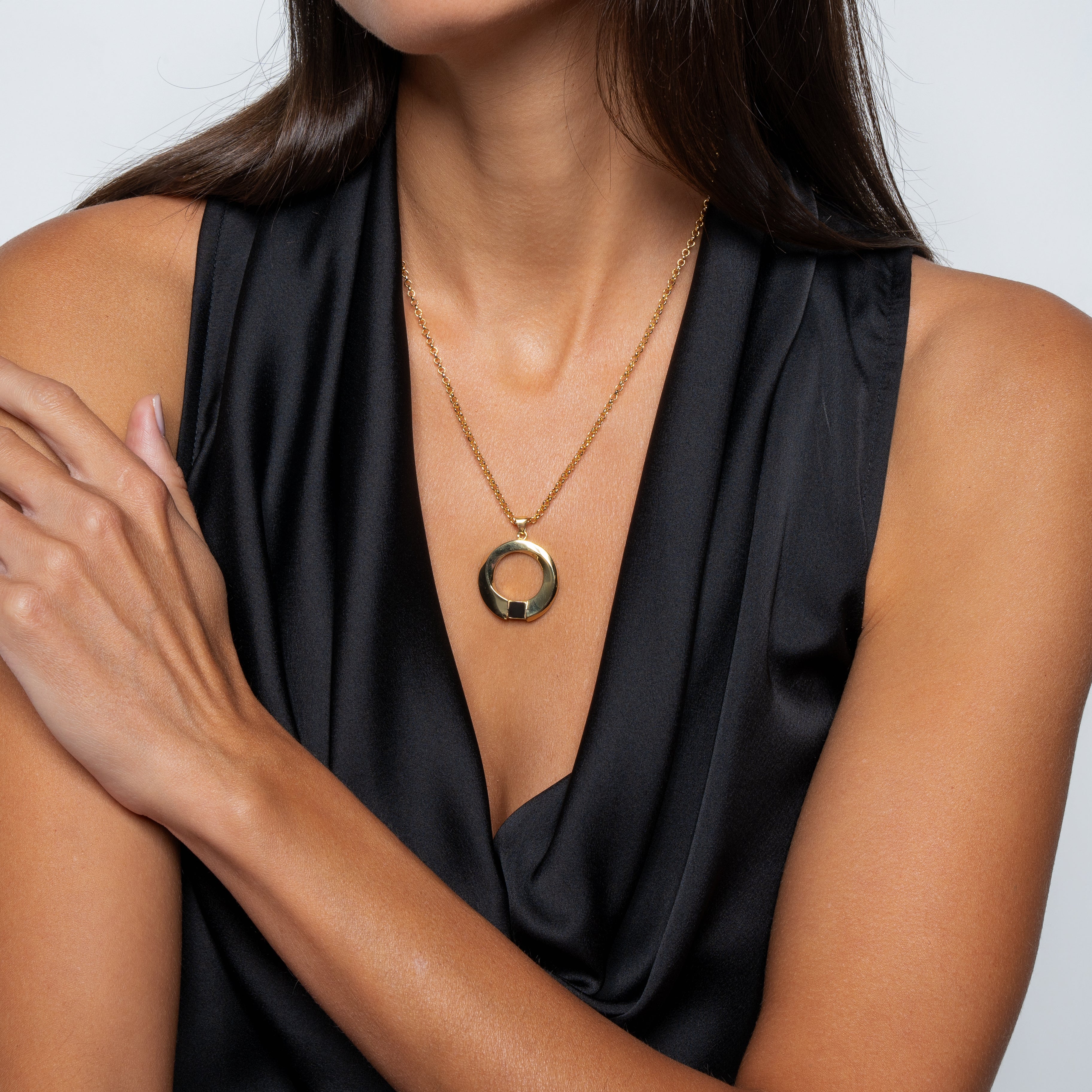 A woman in a sleeveless black top wears the Eternity Circle Necklace, a shiny gold circular pendant. She crosses one arm over her chest, has straight brown hair, and stands against a neutral background.
