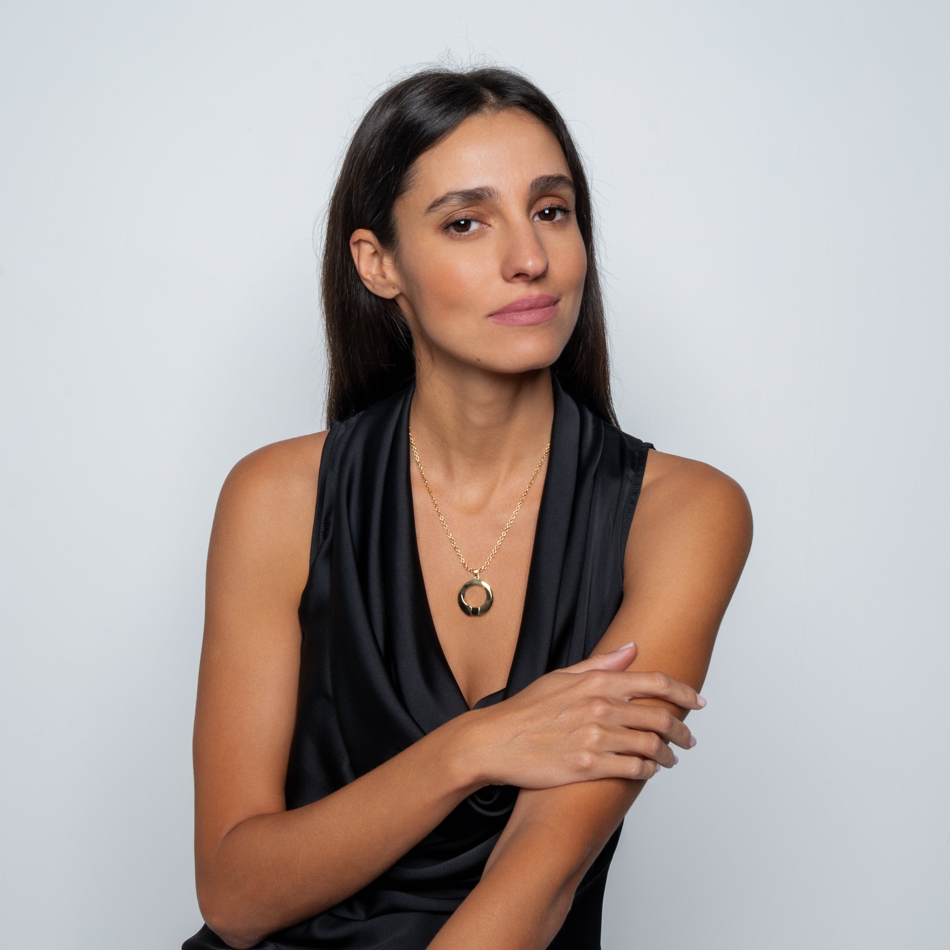 A woman with long brown hair, wearing a sleeveless black top and the Eternity Circle Necklace, sits against a plain light background, resting one arm on the other and looking confidently at the camera.