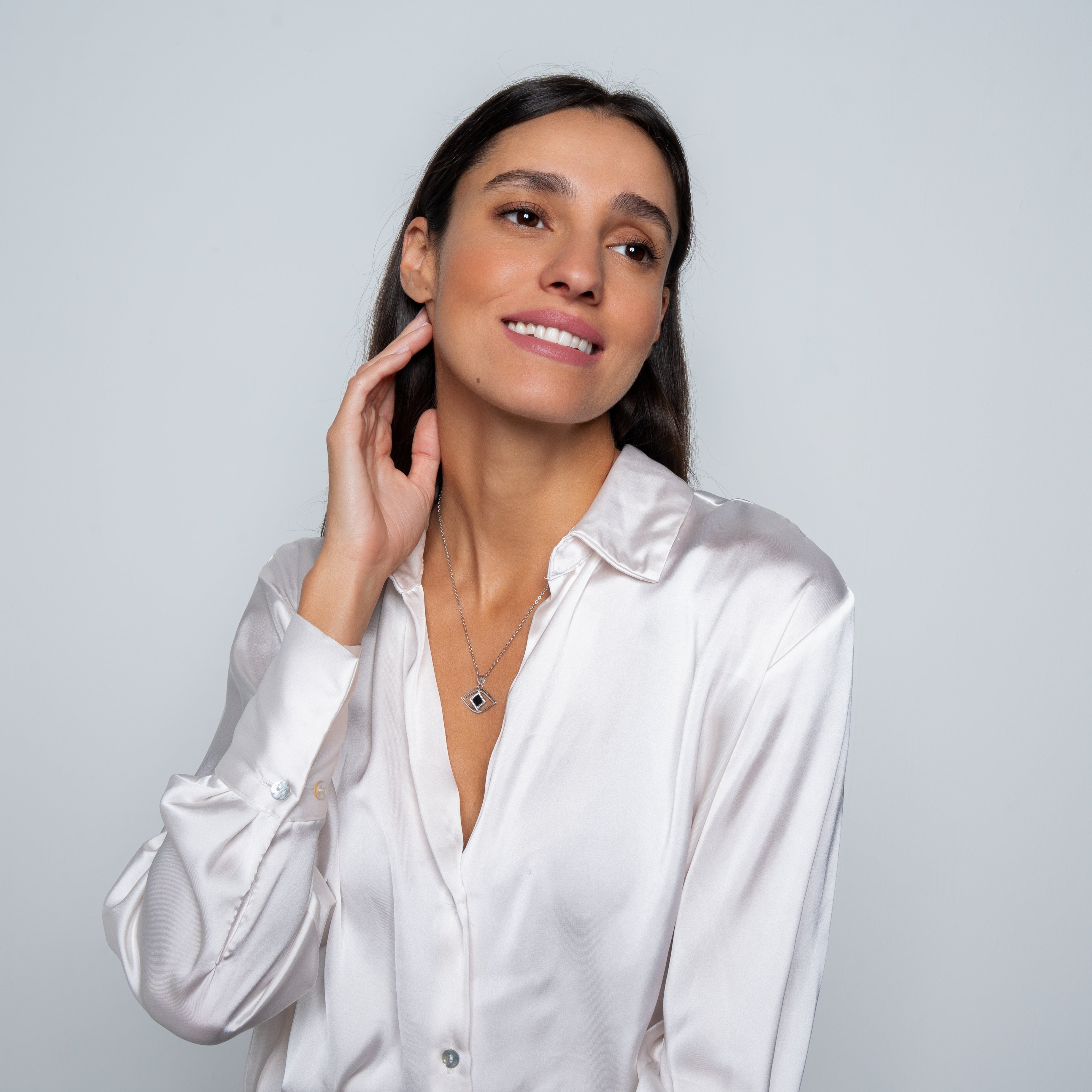 A woman with long dark hair and natural makeup touches her neck, smiling gently as she wears a shiny white satin blouse and the Eye Necklace by Nano Jewelry. The silver necklace features a square nano chip. The background is plain light gray.
