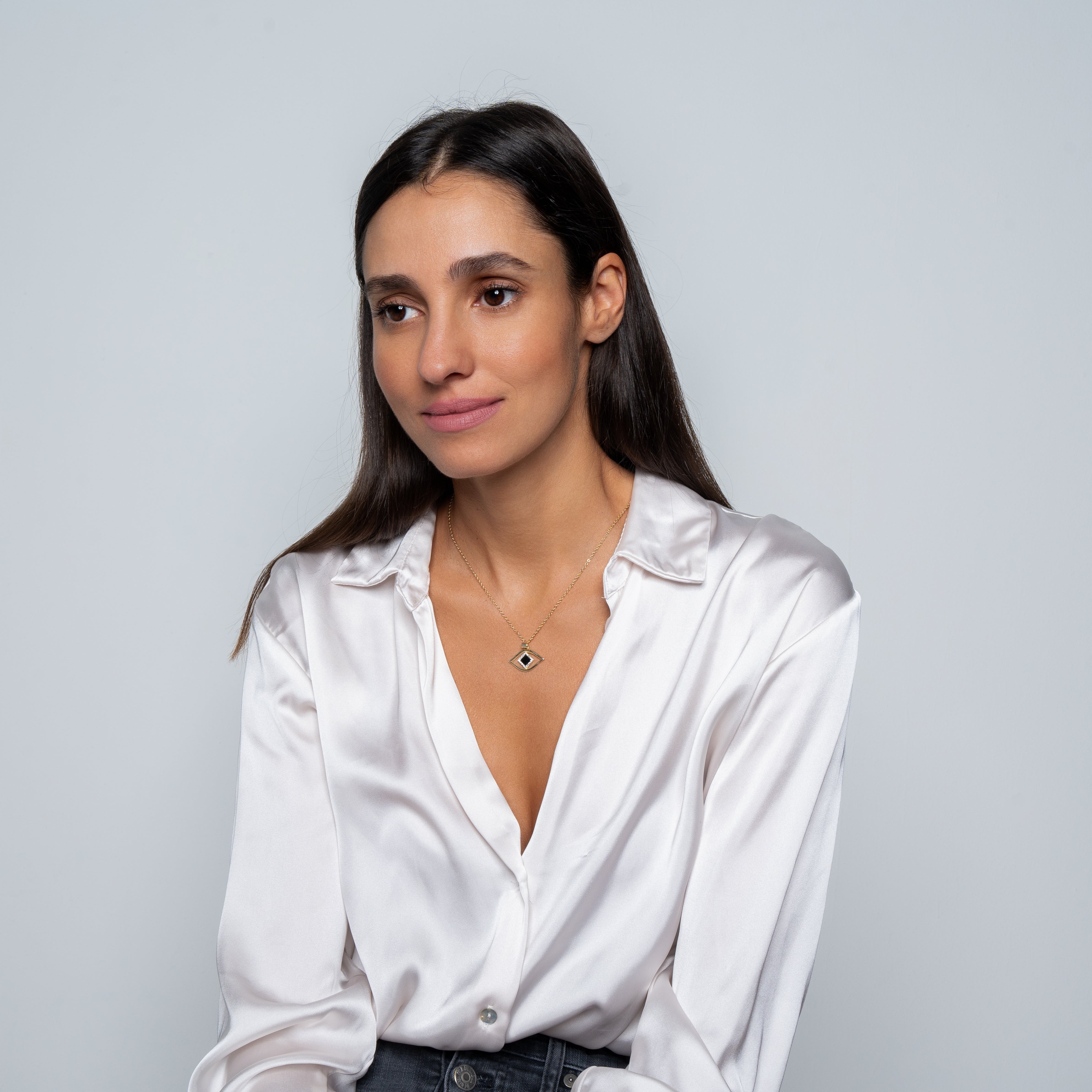 A woman with long brown hair wears a white satin blouse and the Eye Necklace by Nano Jewelry, featuring a geometric pendant, while sitting against a plain light gray background and gazing calmly to the side.