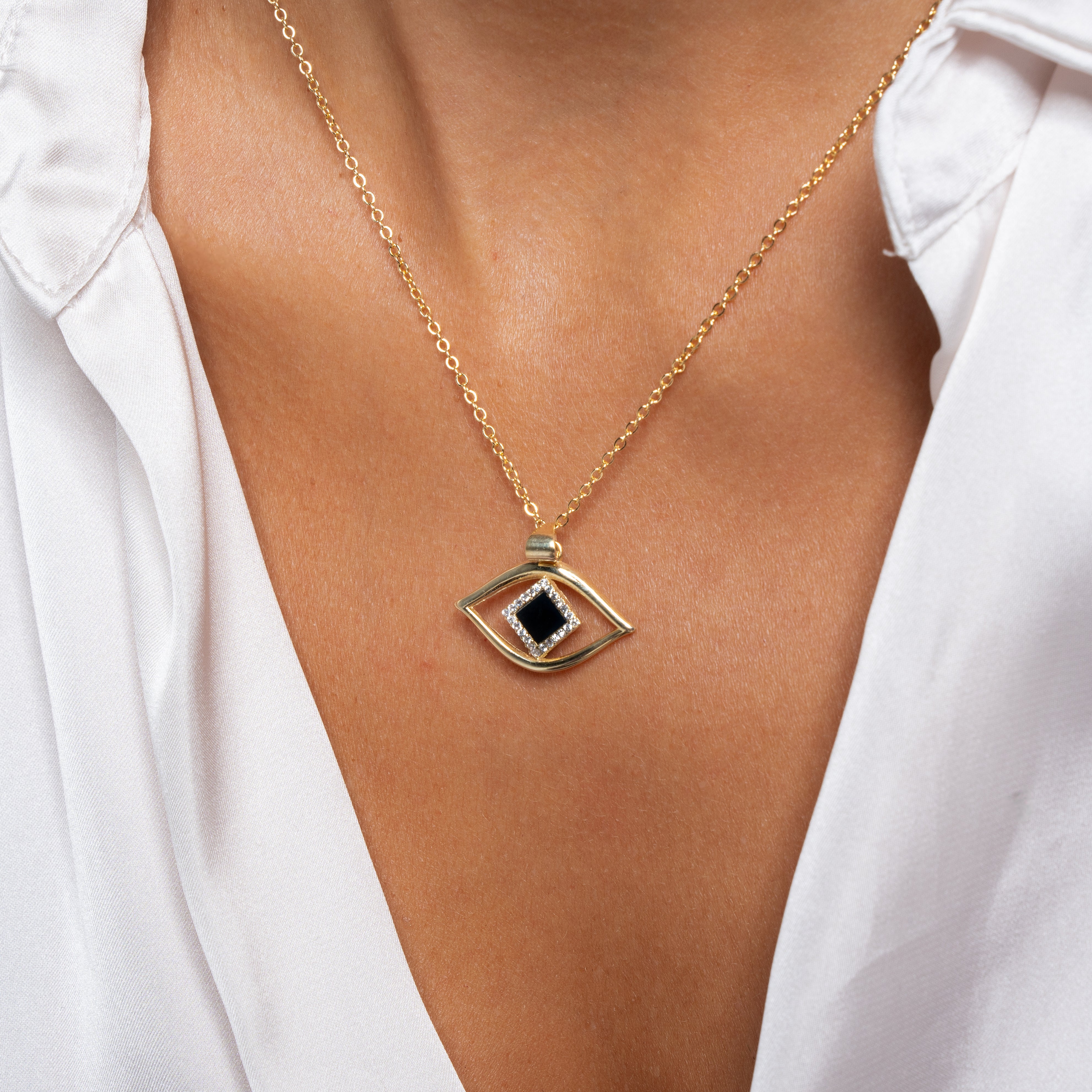 A person wears a white shirt with the Eye Necklace, a gold nano jewelry piece featuring an eye-shaped pendant with a black center stone, resting on their chest.
