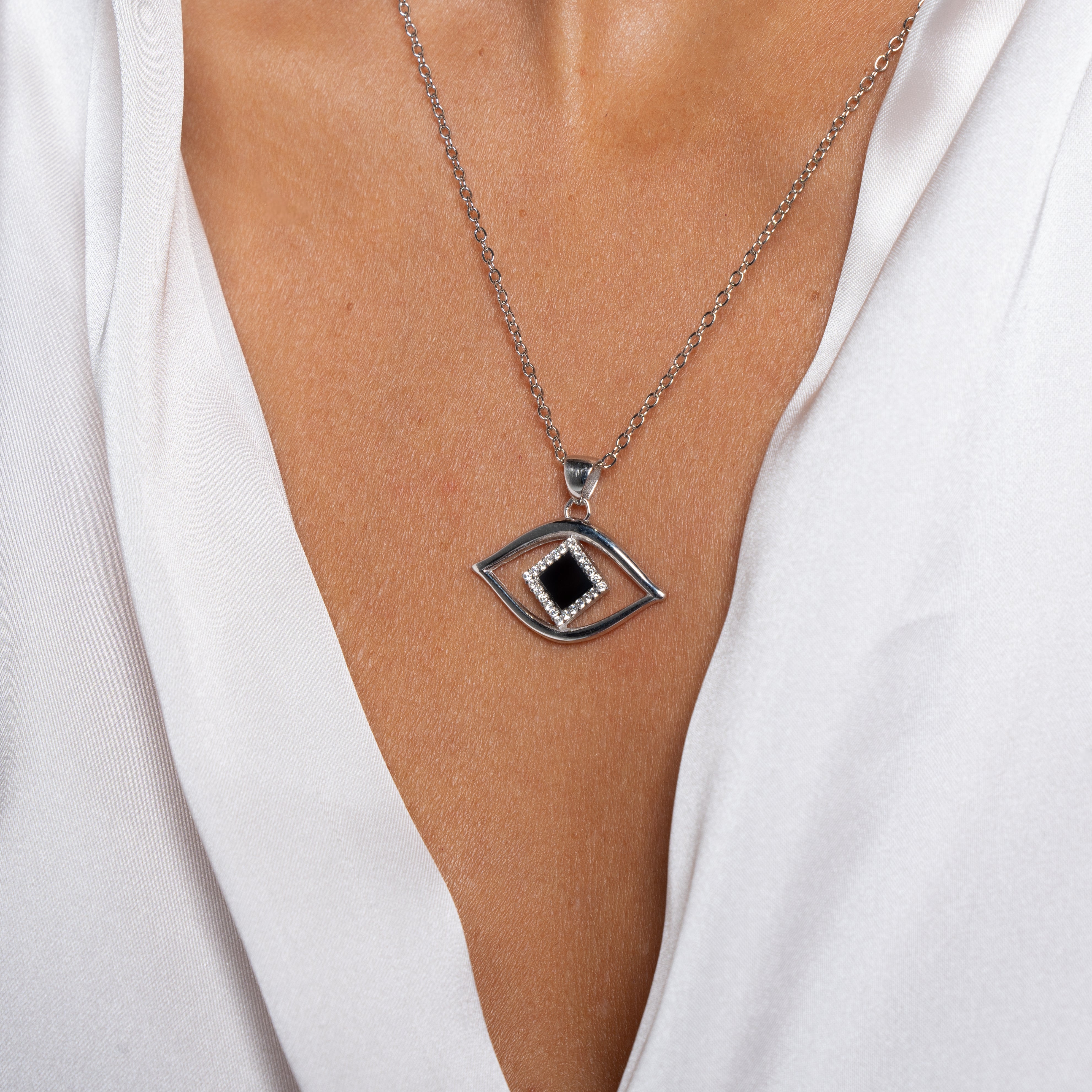 A person wearing a white V-neck blouse displays the Eye Necklace, a silver Nano Jewelry pendant with an eye shape, featuring a square black nano chip and small clear gems, on their chest.