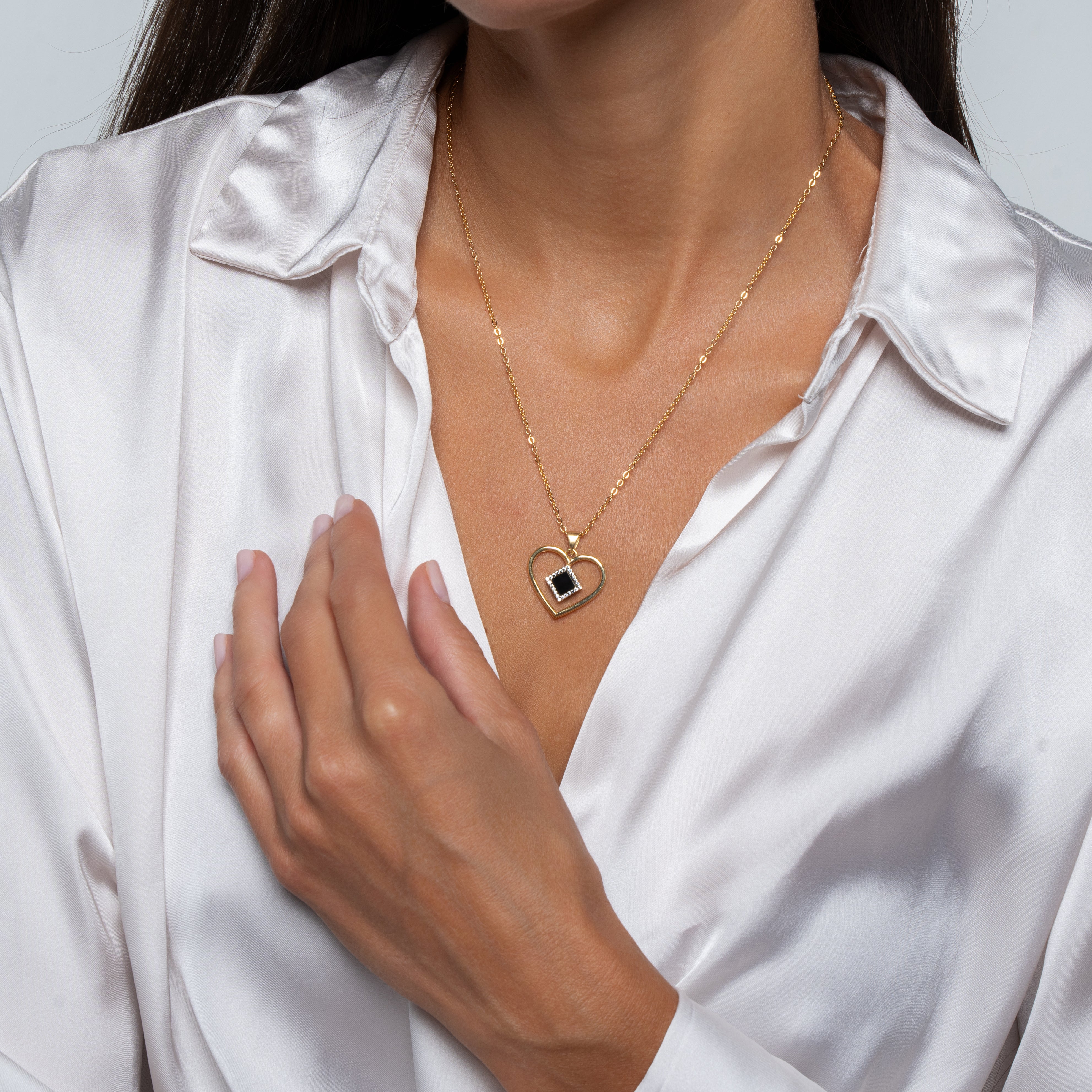 A woman in a white satin blouse adjusts her collar, showcasing the gold Heart Necklace | Old Testament Edition from NanoBless—an elegant piece of Jewish jewelry featuring a black stone heart-shaped pendant.