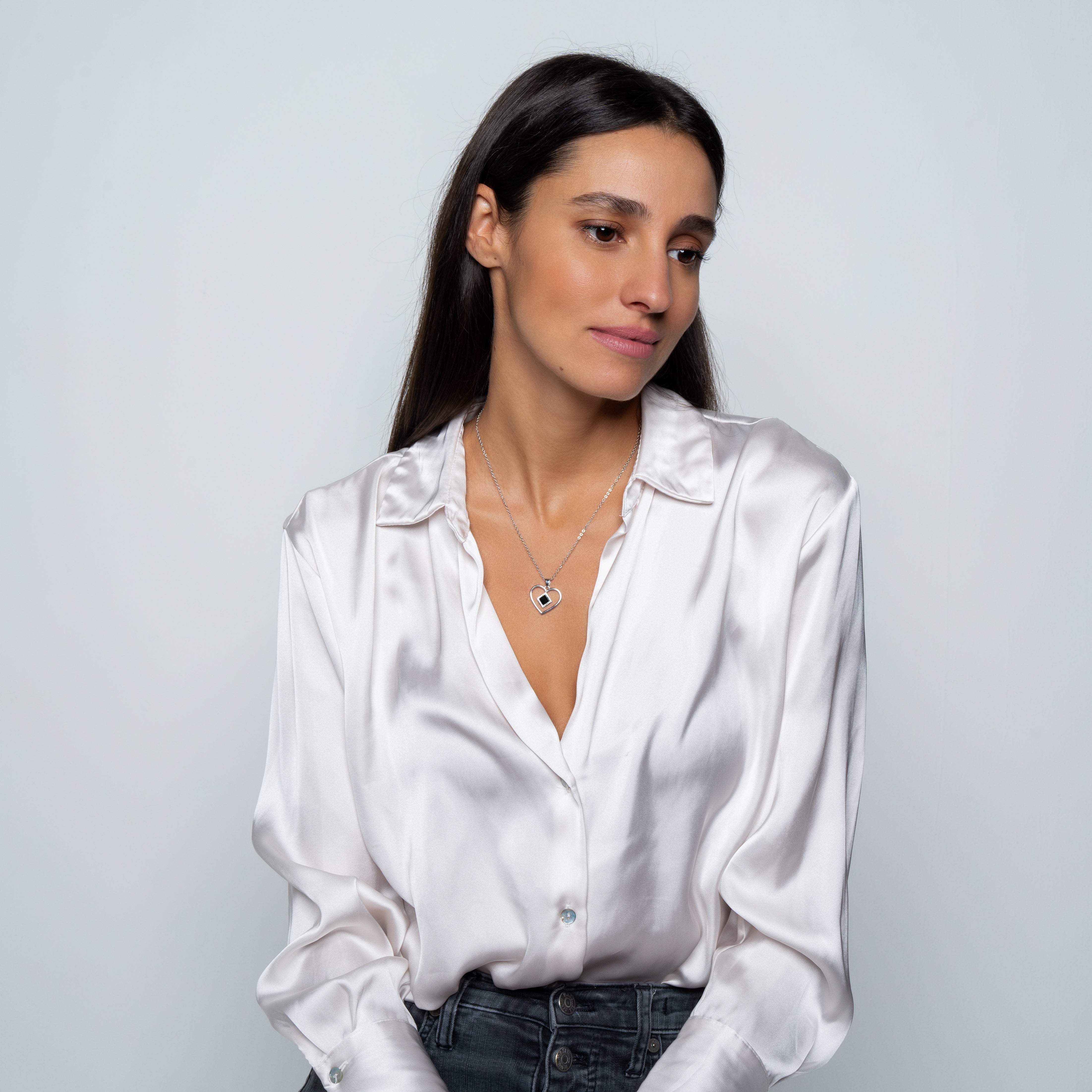A woman with long dark hair, wearing a shiny white satin blouse and the Heart Necklace | Old Testament Edition, gazes gently to the side against a plain light gray background.