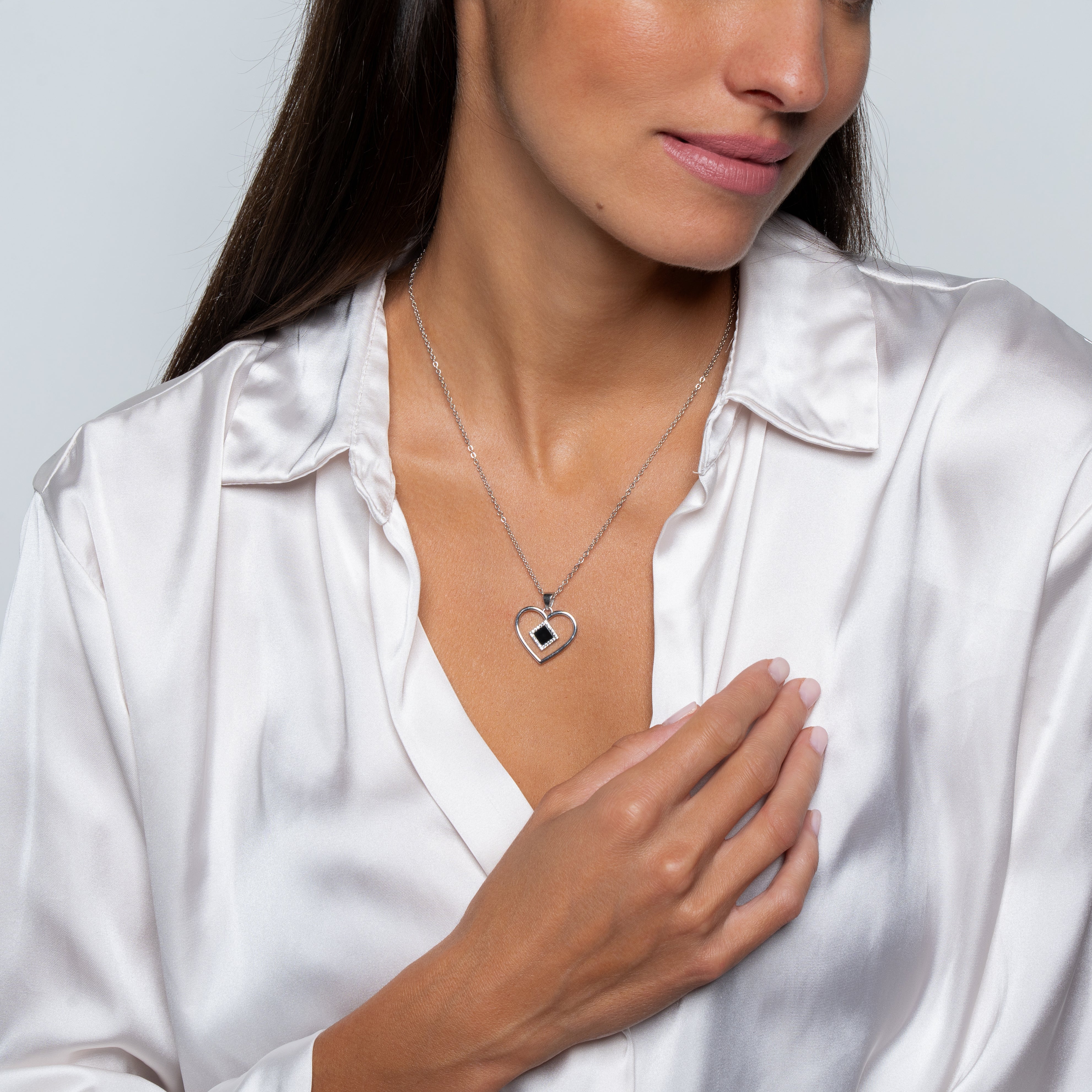 A woman with long dark hair smiles softly, touching her chest to showcase the Heart Necklace | Old Testament Edition— a silver heart-shaped pendant—while wearing a silky white blouse against a plain, light background.