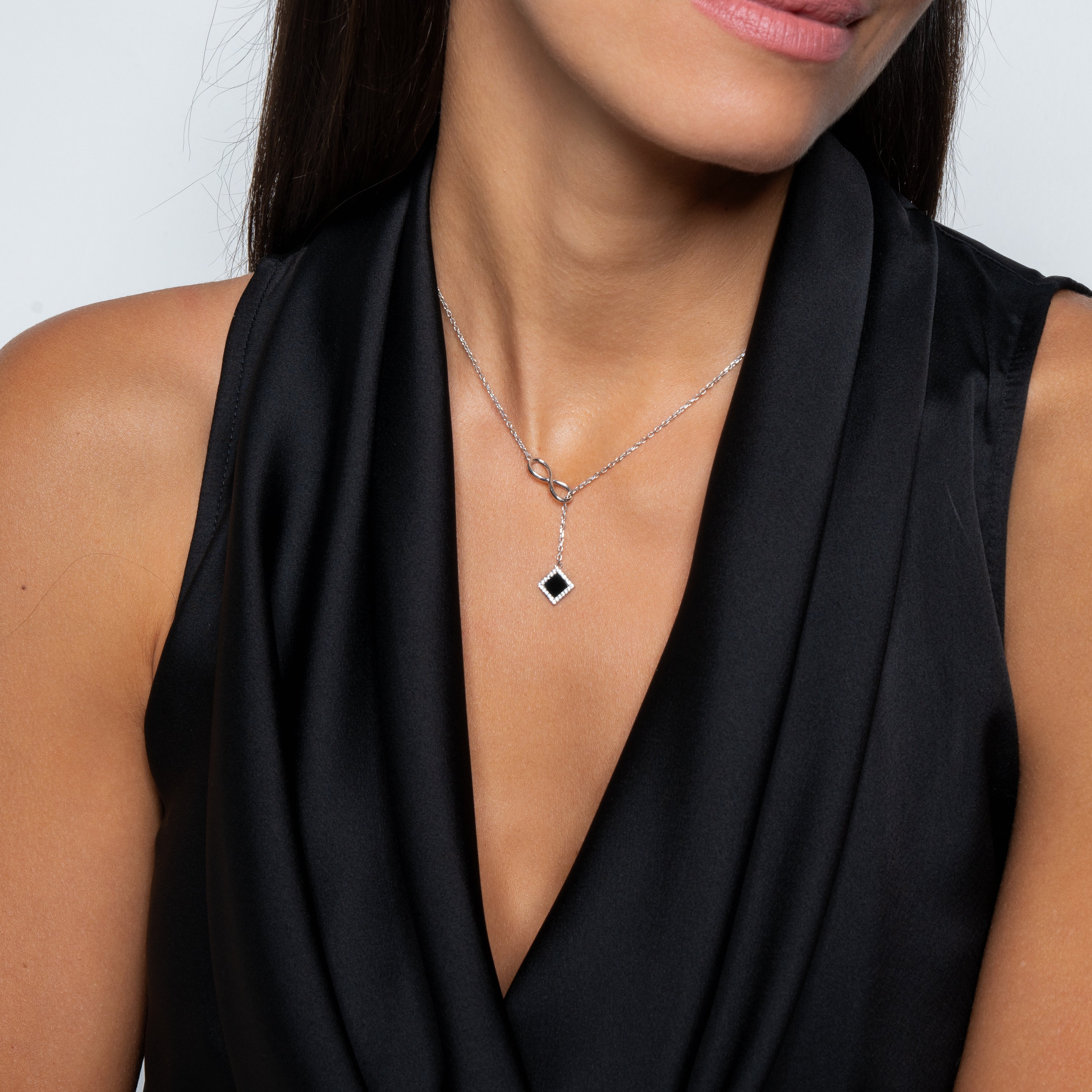 A woman in a sleeveless black dress showcases the Infinity Necklace - Old Testament Edition, featuring a silver chain with intricate Jewish Jewelry details and a black diamond-shaped pendant. The focus is on her neck and upper chest; her face is partially visible.