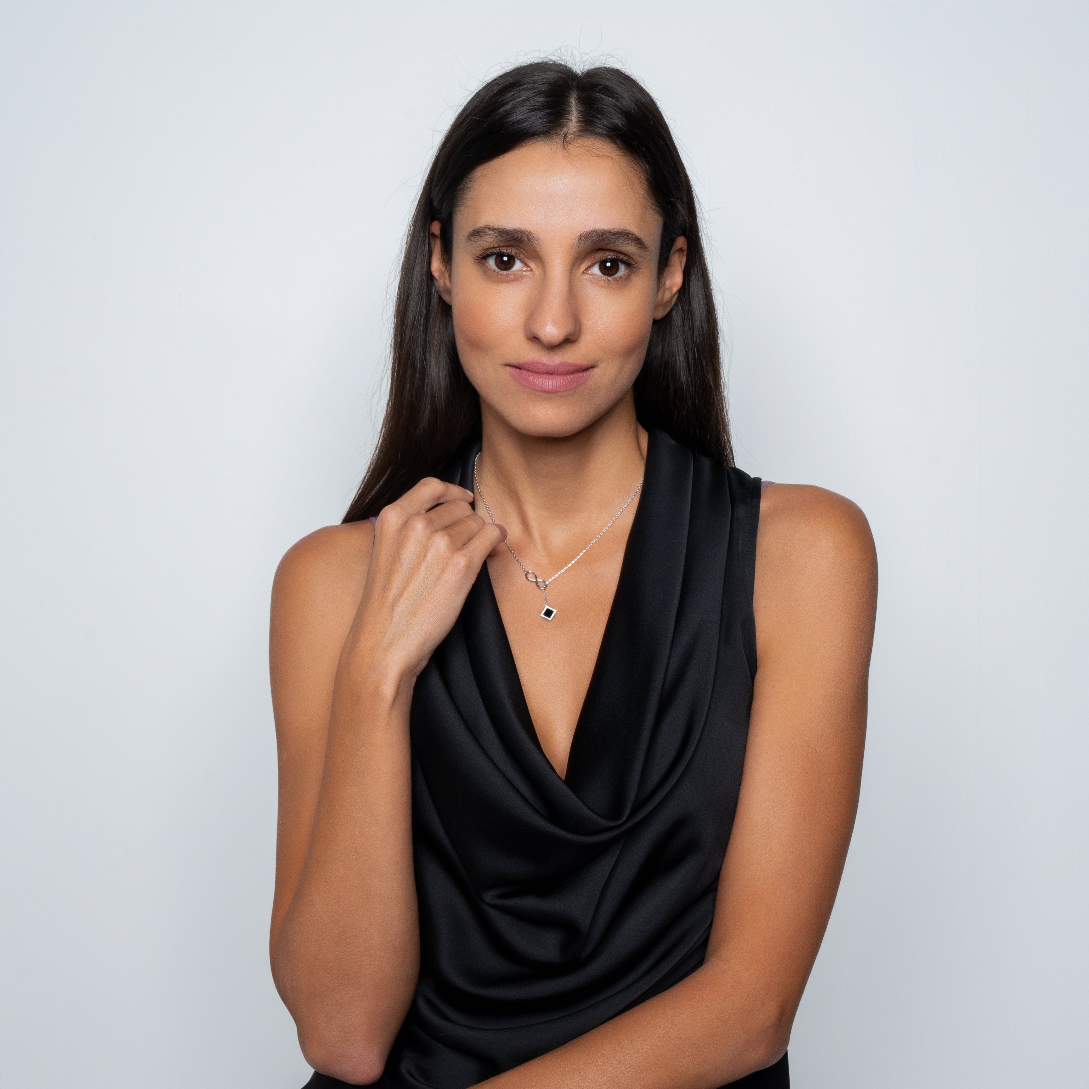 A woman with long dark hair, in a sleeveless black satin top, poses against a light background. She holds the Infinity Necklace- Old Testament Edition with a small pendant, gazing calmly at the camera.