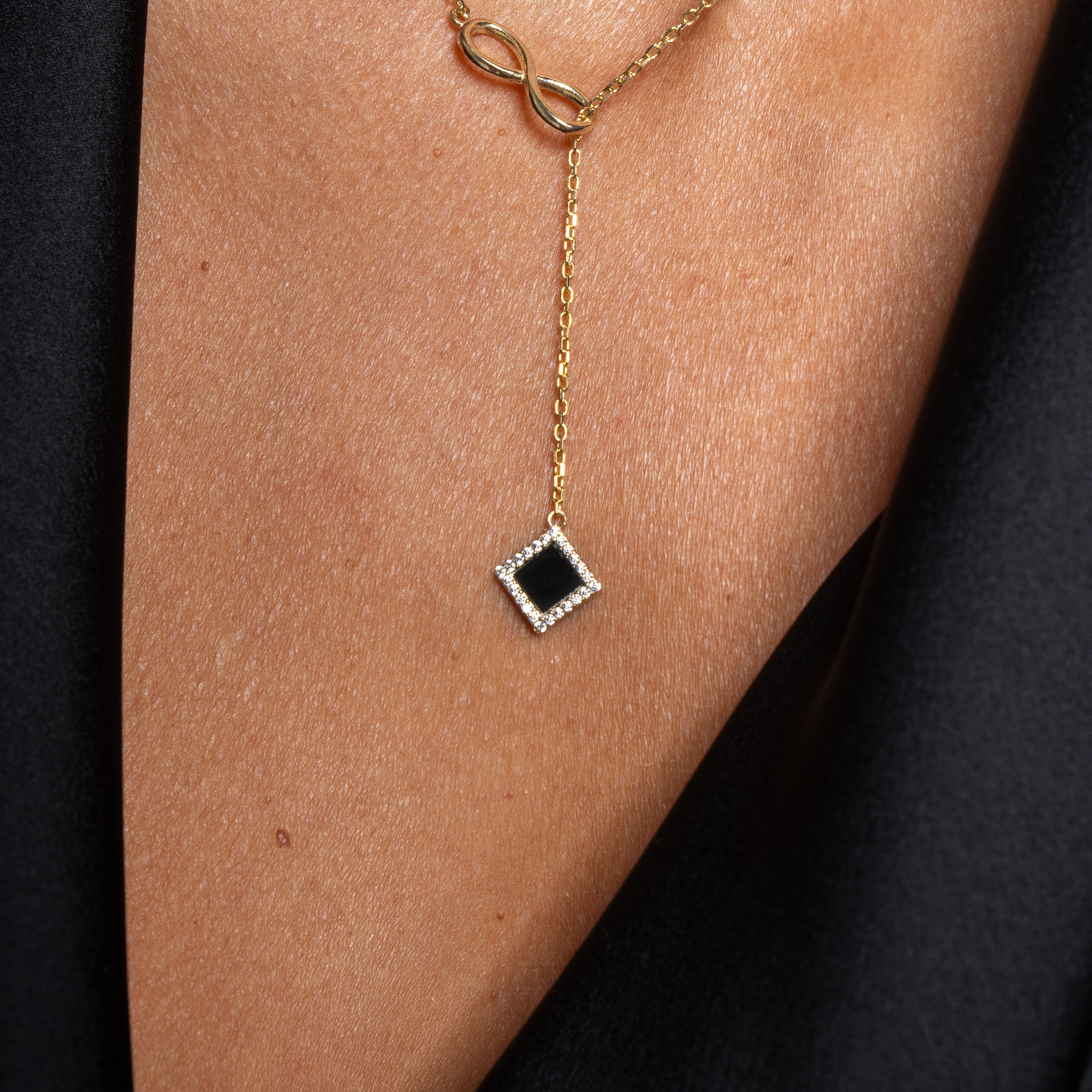 A close-up of the Infinity Necklace–Old Testament Edition: a gold necklace featuring an infinity symbol and a square black pendant with clear stone accents, worn on bare skin and partially covered by black fabric—a stunning Nano Jewelry piece.