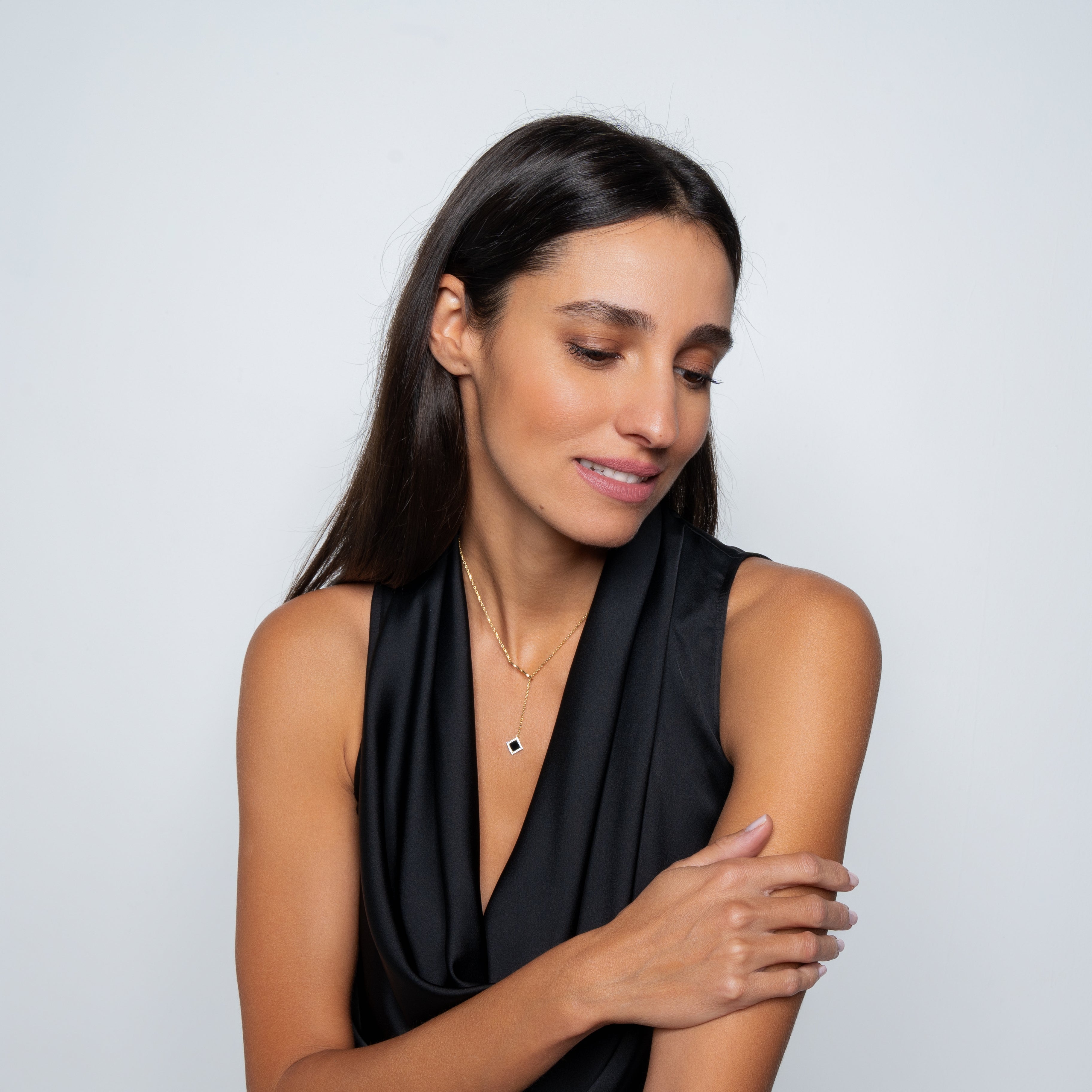 A woman with long dark hair wears a sleeveless black top and the Infinity Necklace- Old Testament Edition from Nano Jewelry, featuring a gold chain and black pendant, as she smiles gently, looking down and touching her arm against a plain light background.