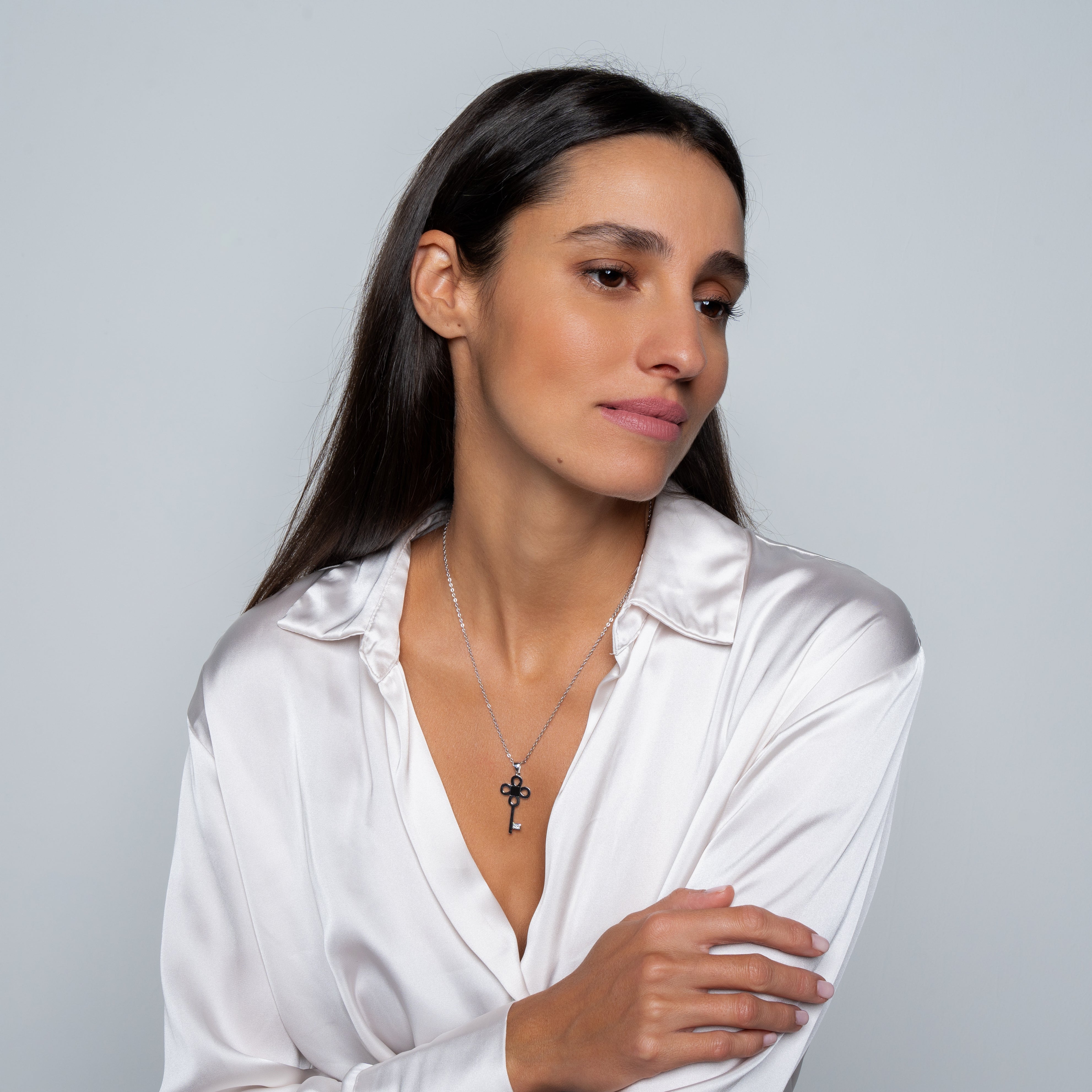 A woman with long dark hair in a white satin blouse wears the Key Necklace- Old Testament Edition, posing against a plain light gray background and gazing calmly to the side.