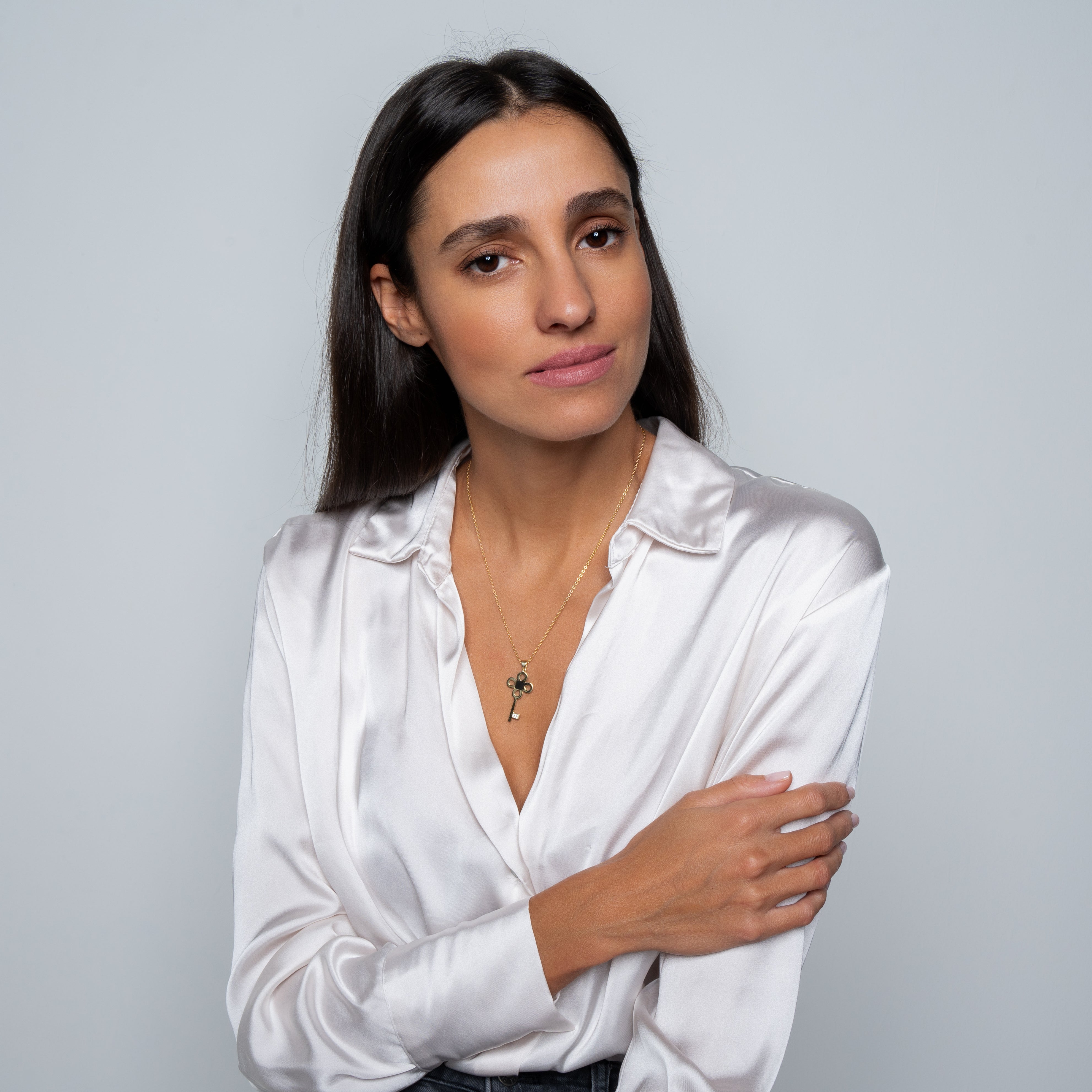 A woman with long dark hair, in a satin white blouse and the Key Necklace—Old Testament Edition, an elegant nano jewelry piece, poses cross-armed and expressionless against a plain light background.