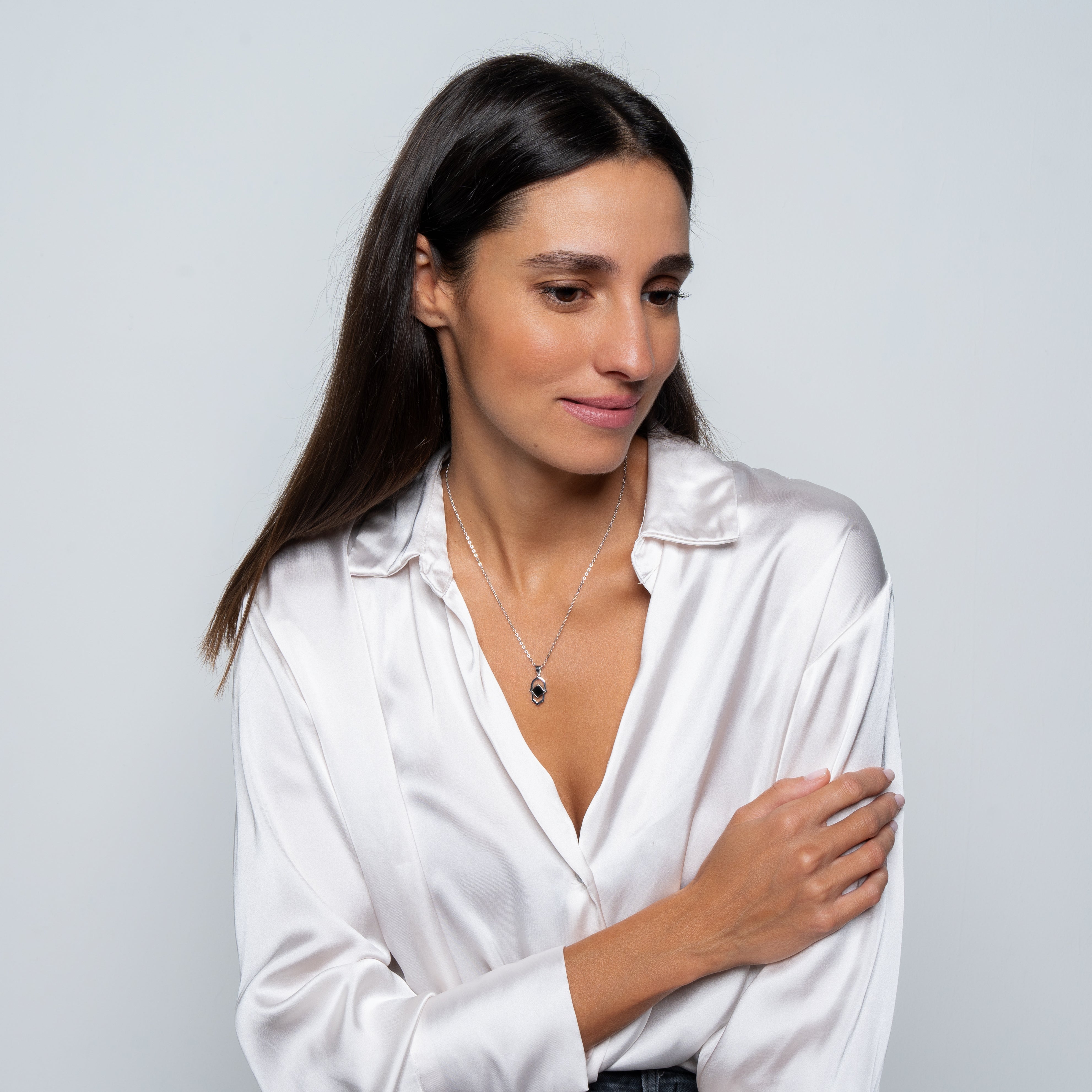 A woman with long brown hair, dressed in a white satin blouse and wearing the Basic Hamsa silver necklace with a  pendant, stands against a light gray background, looking down gently.