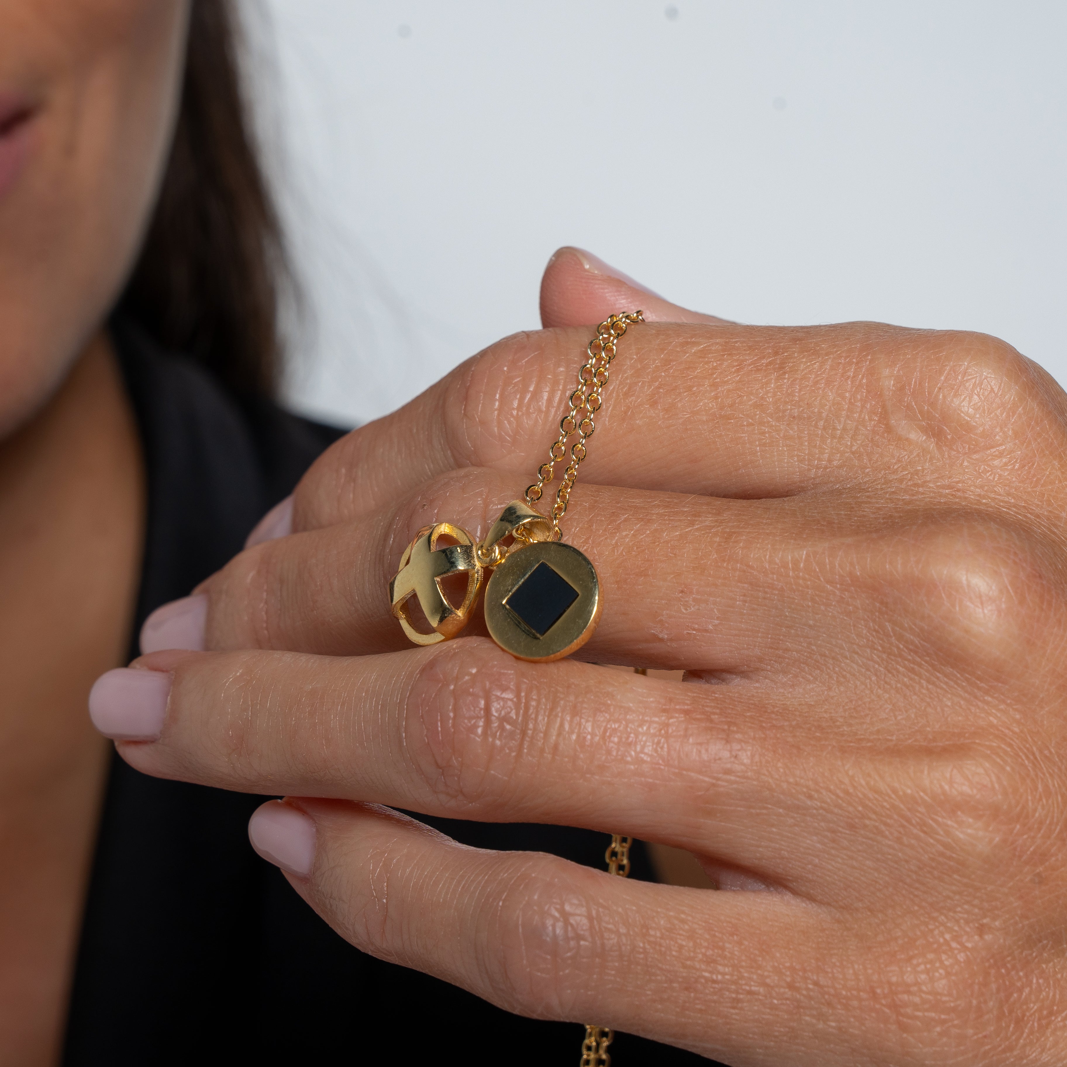 A person with light pink nails holds the Opening Cross necklace, a gold Christian Jewelry piece with a round pendant featuring a black geometric design. Part of their smiling face is visible on the left, set against a plain light background.