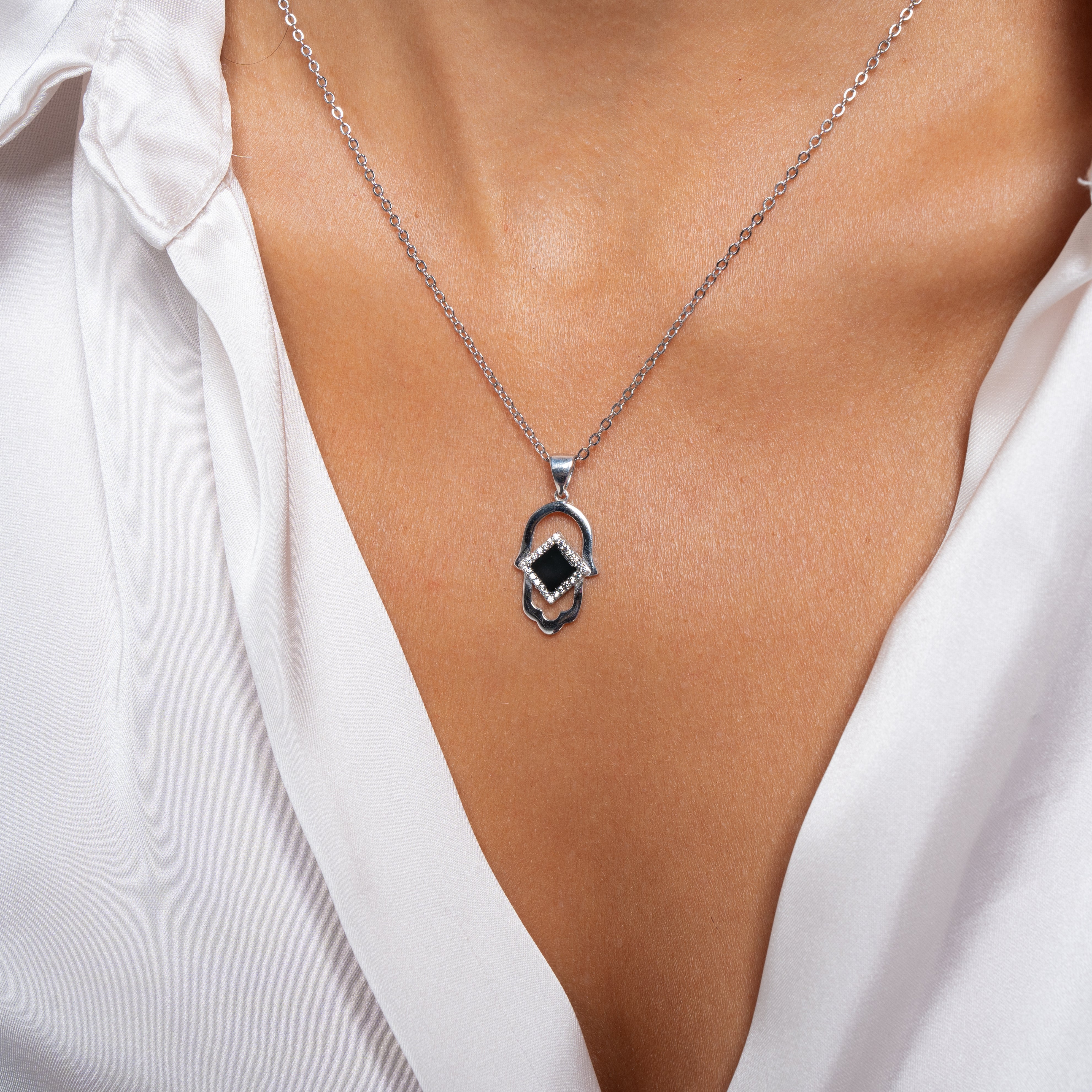 A close-up of a person wearing the Hamsa Necklace | Old Testament Edition by Nano Jewelry, featuring a silver chain and Hamsa pendant with a black center stone, styled over a white, partially unbuttoned shirt.