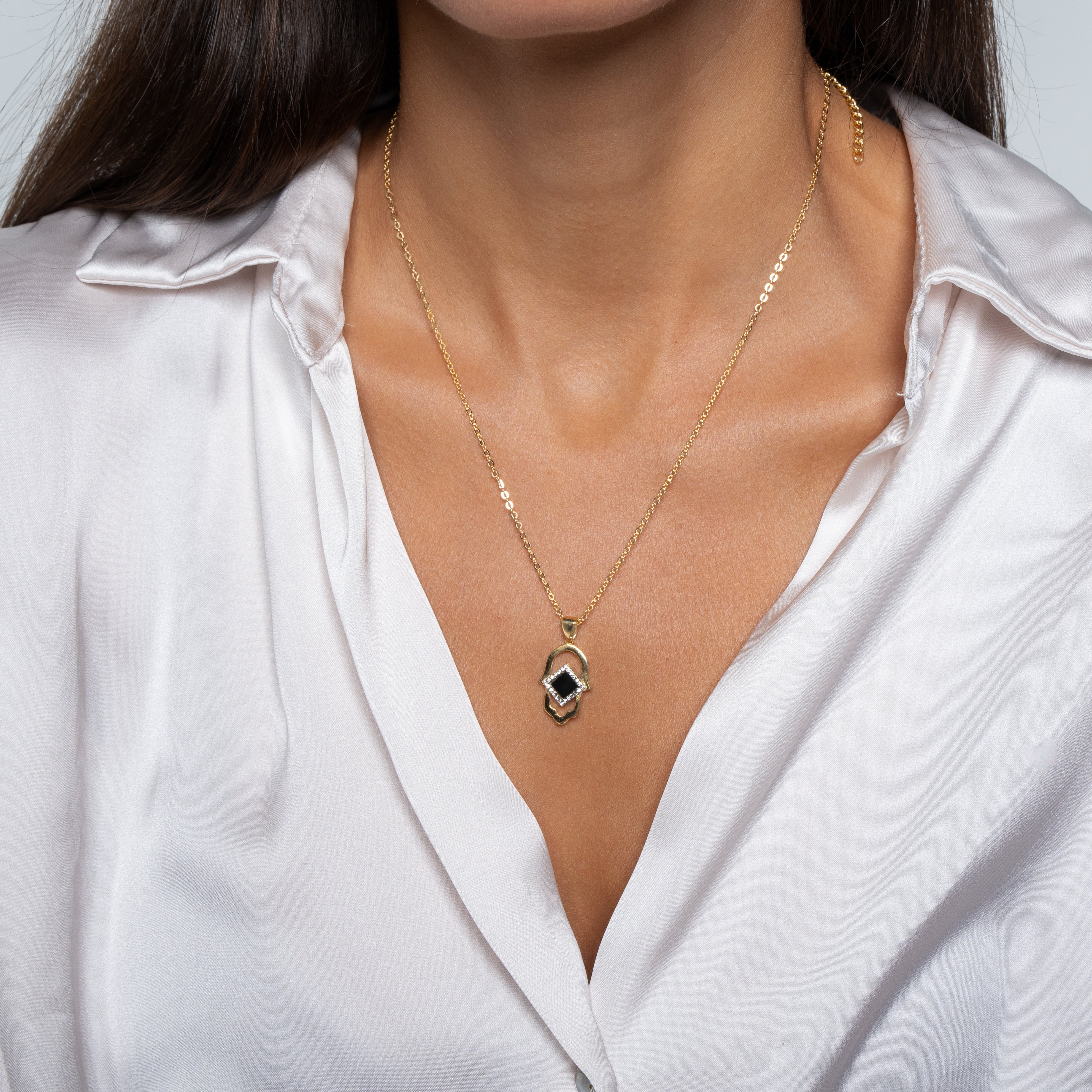 A woman in a white satin blouse models the Hamsa Necklace | Old Testament Edition—a gold Christian jewelry piece with a geometric, black-and-gold pendant resting at the center of her chest.