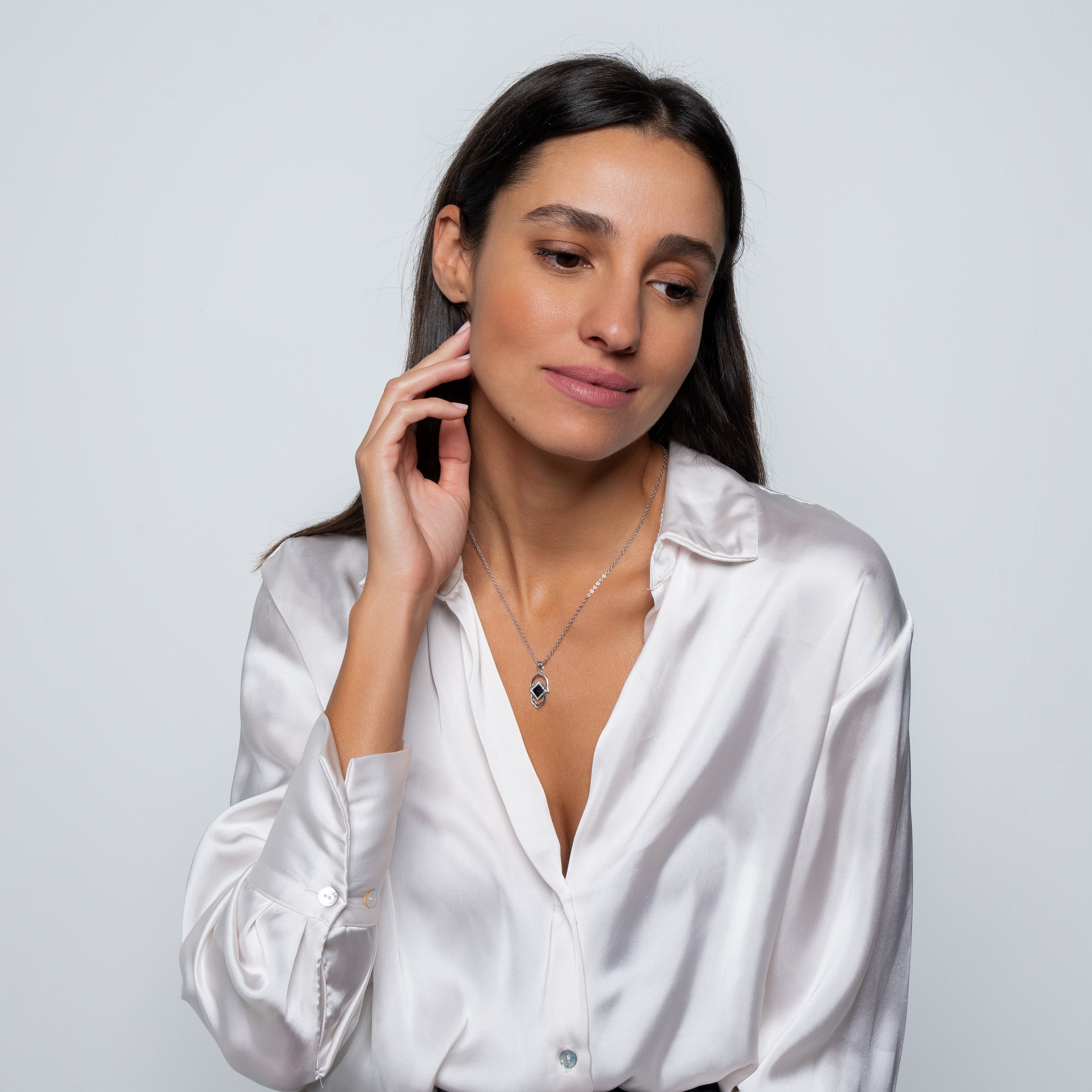 Woman with long dark hair wearing a silky white blouse and the Hamsa Necklace | Old Testament Edition, posing with one hand touching her neck and looking down to the side against a plain light background.