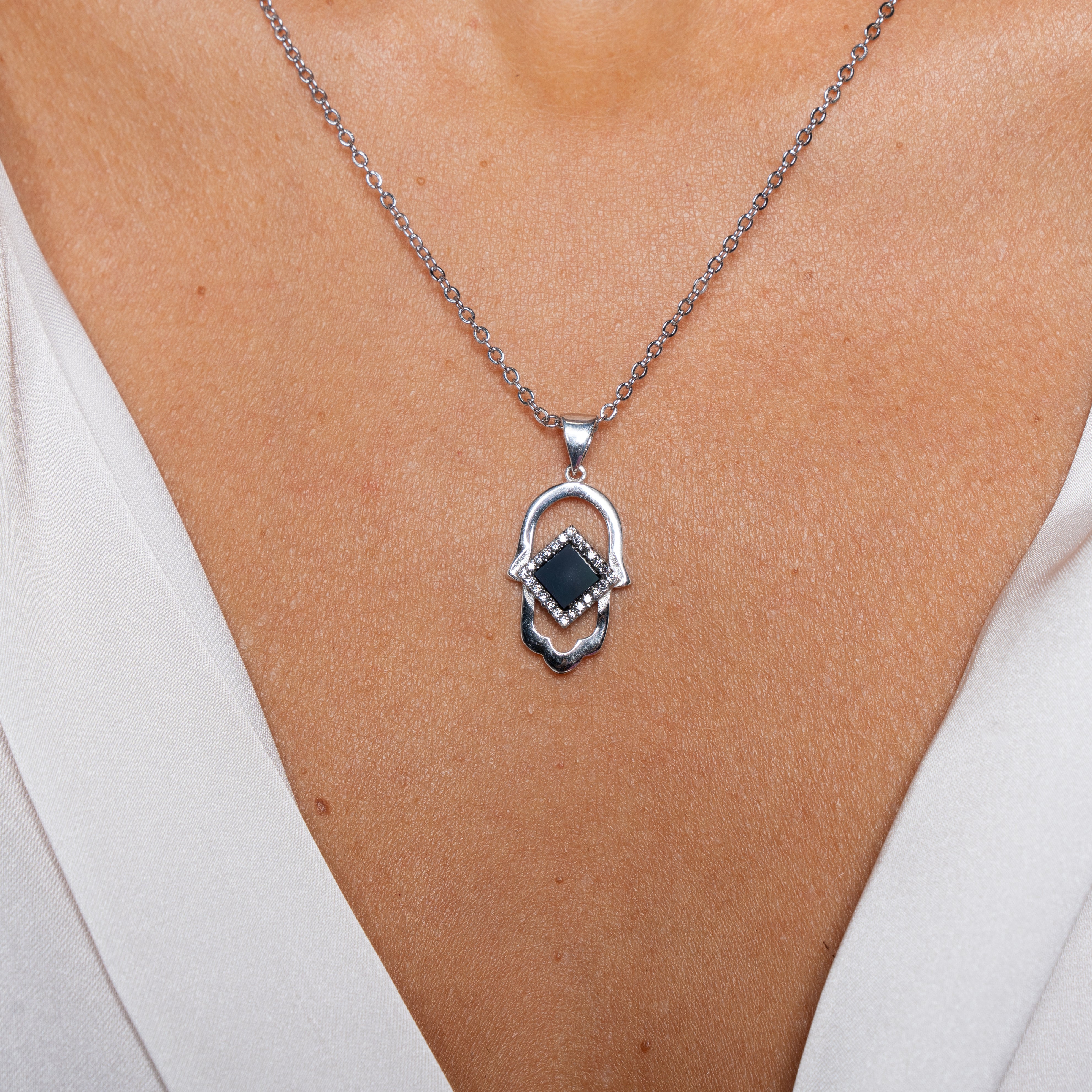 A close-up of someone wearing the Hamsa Necklace | Old Testament Edition, a silver piece with a black stone Hamsa hand pendant, shown against bare skin and a white V-neck top.