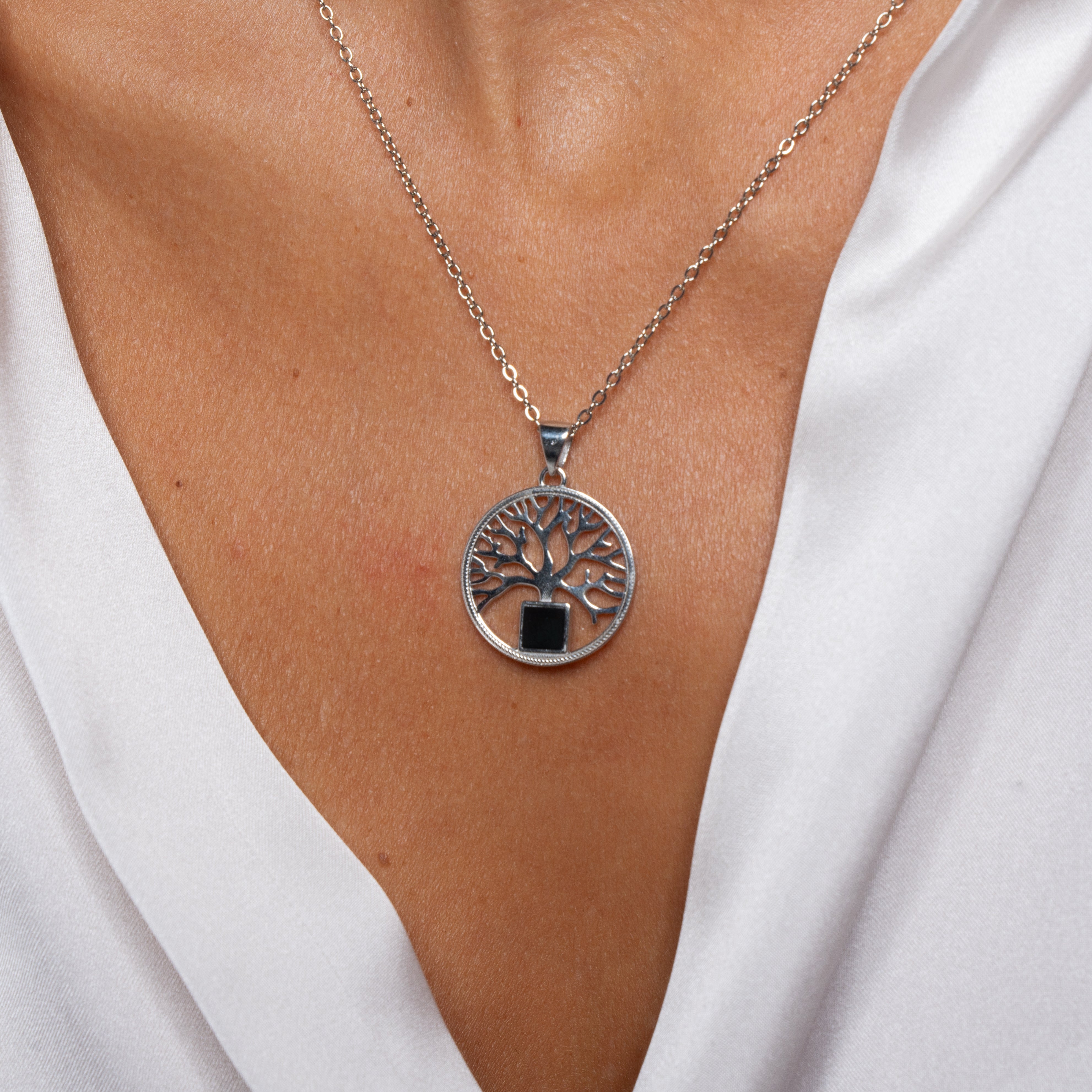 A close-up of a person wearing the Tree of Life pendant necklace by NanoBless over a white V-neck blouse; the silver pendant features an intricate tree design with a small black rectangle at its base.