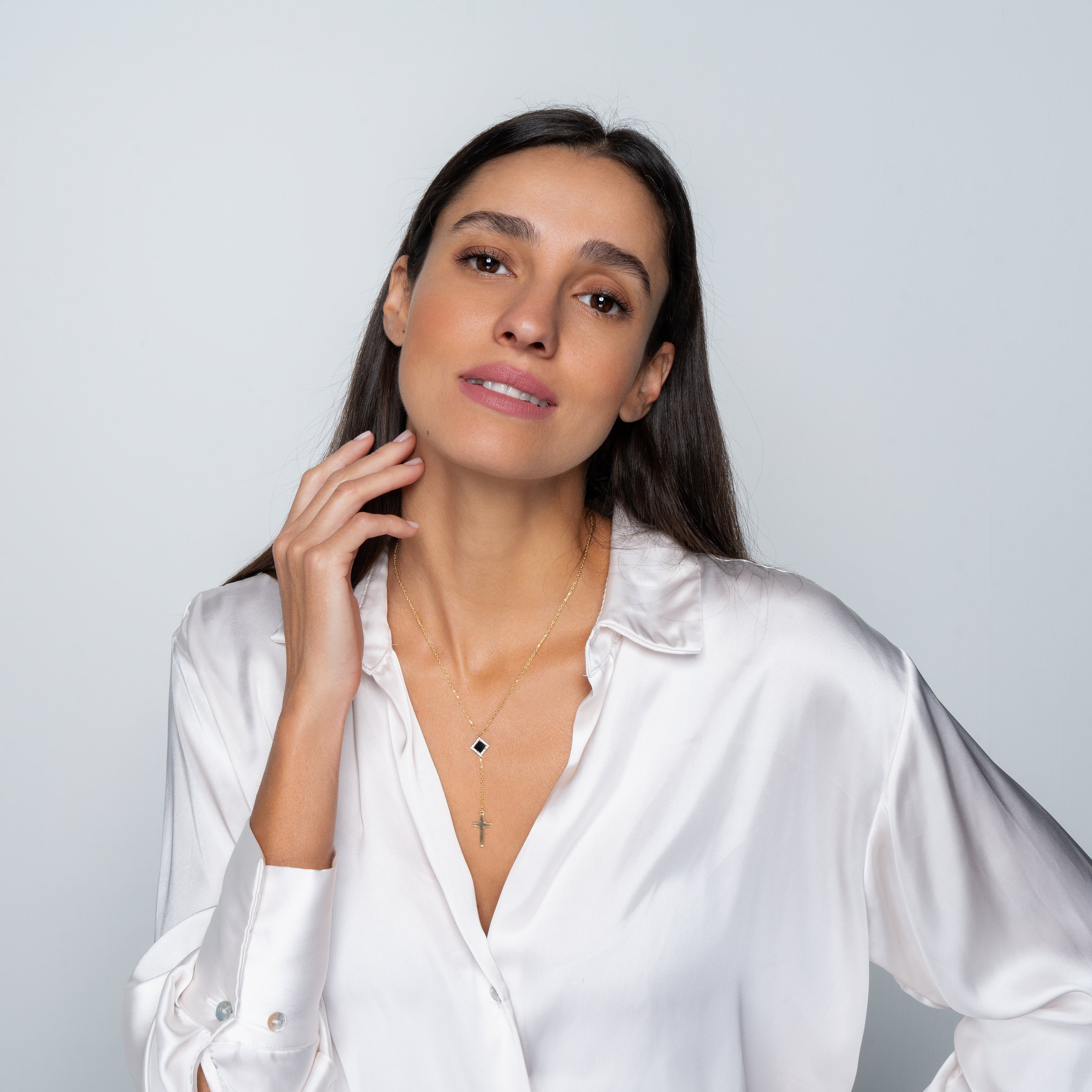 A woman with long dark hair wears a shiny white blouse and a layered Mini Cross Y Necklace, standing against a light background. She smiles gently with one hand touching her face.