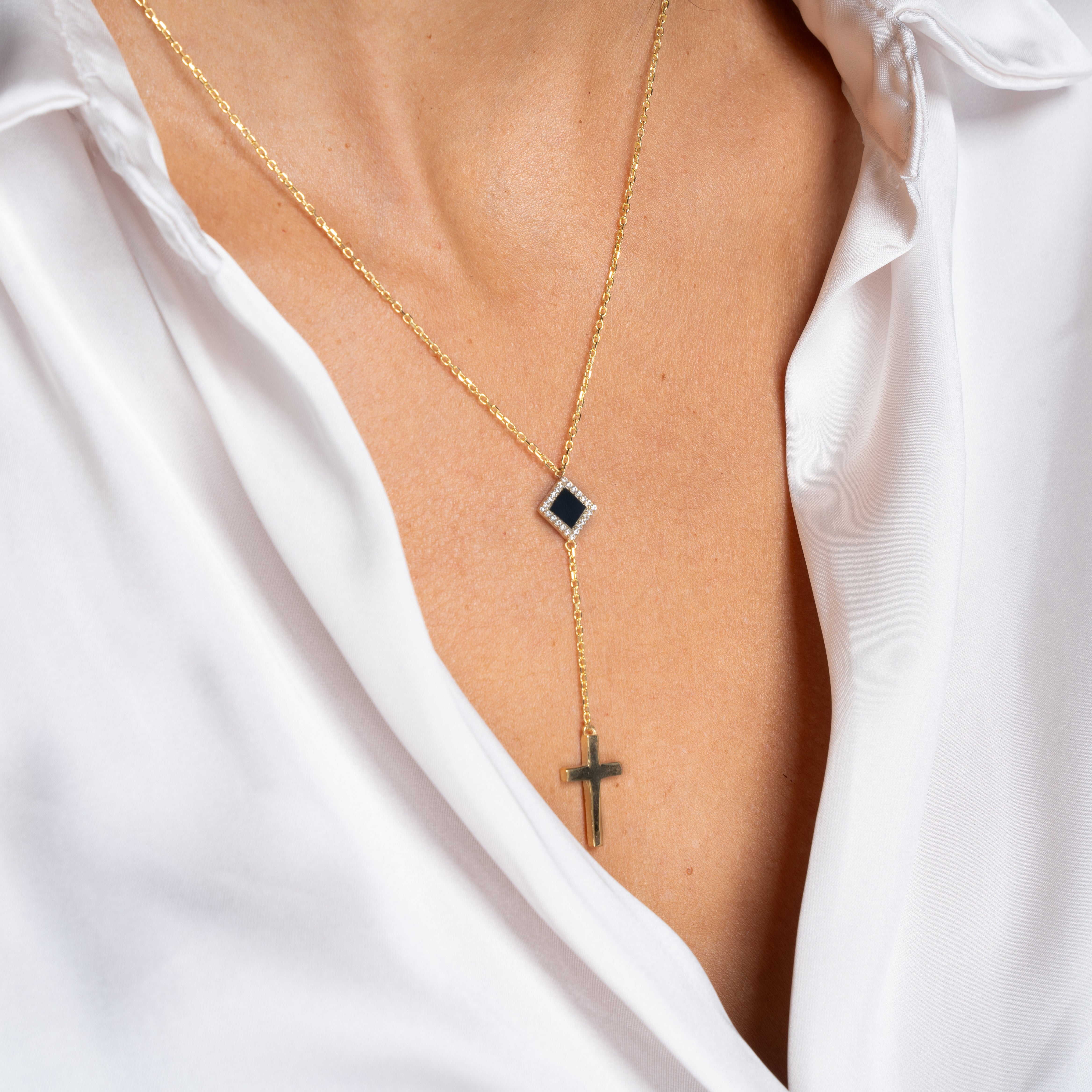 A person wearing a white shirt with an open collar, showcasing Christian Jewelry: the Mini Cross Y Necklace, a gold piece featuring a black square pendant and a hanging gold cross.