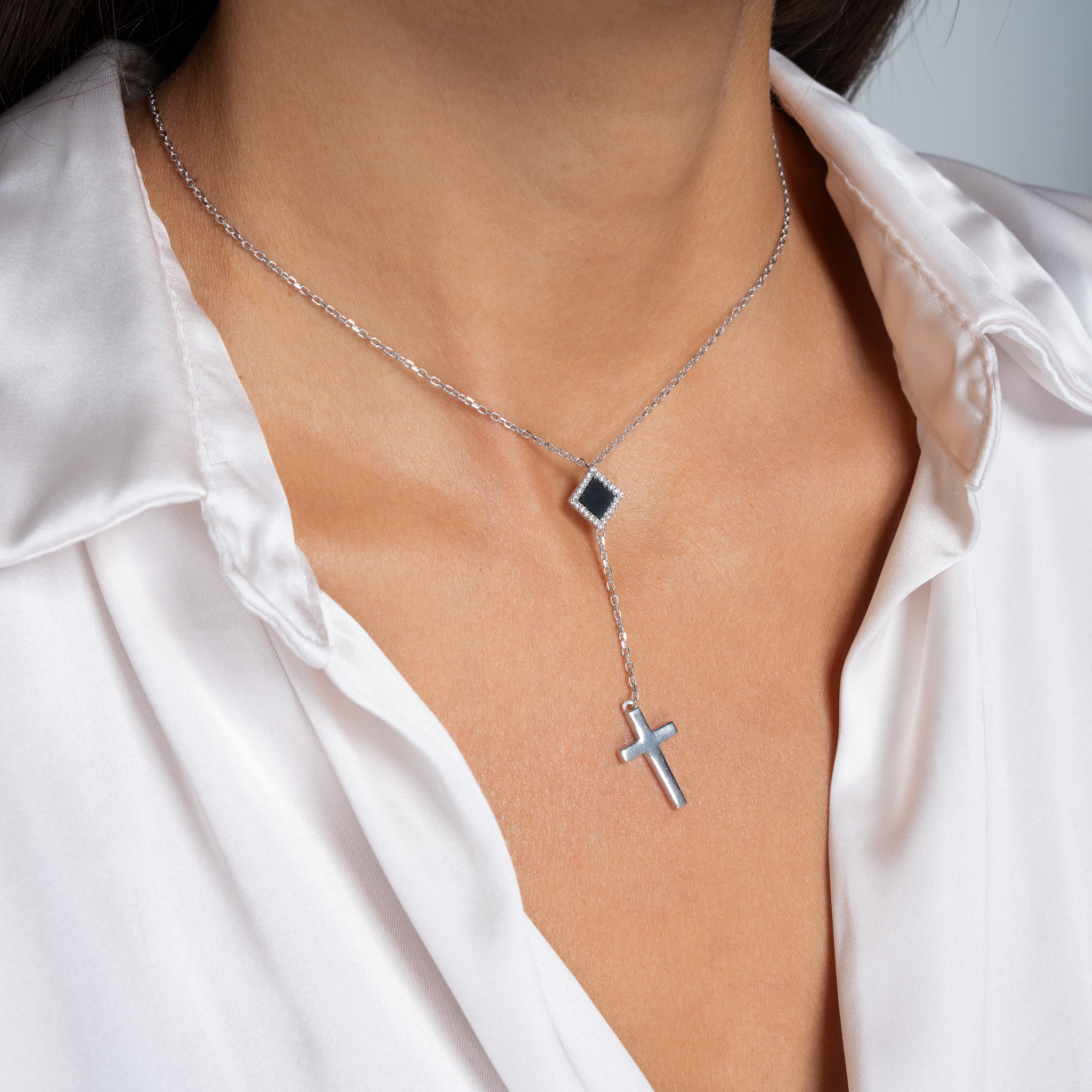 A close-up of a woman wearing the Mini Cross Y Necklace—a delicate silver piece with a black square pendant and a dangling cross—paired with an open-collar white satin blouse.