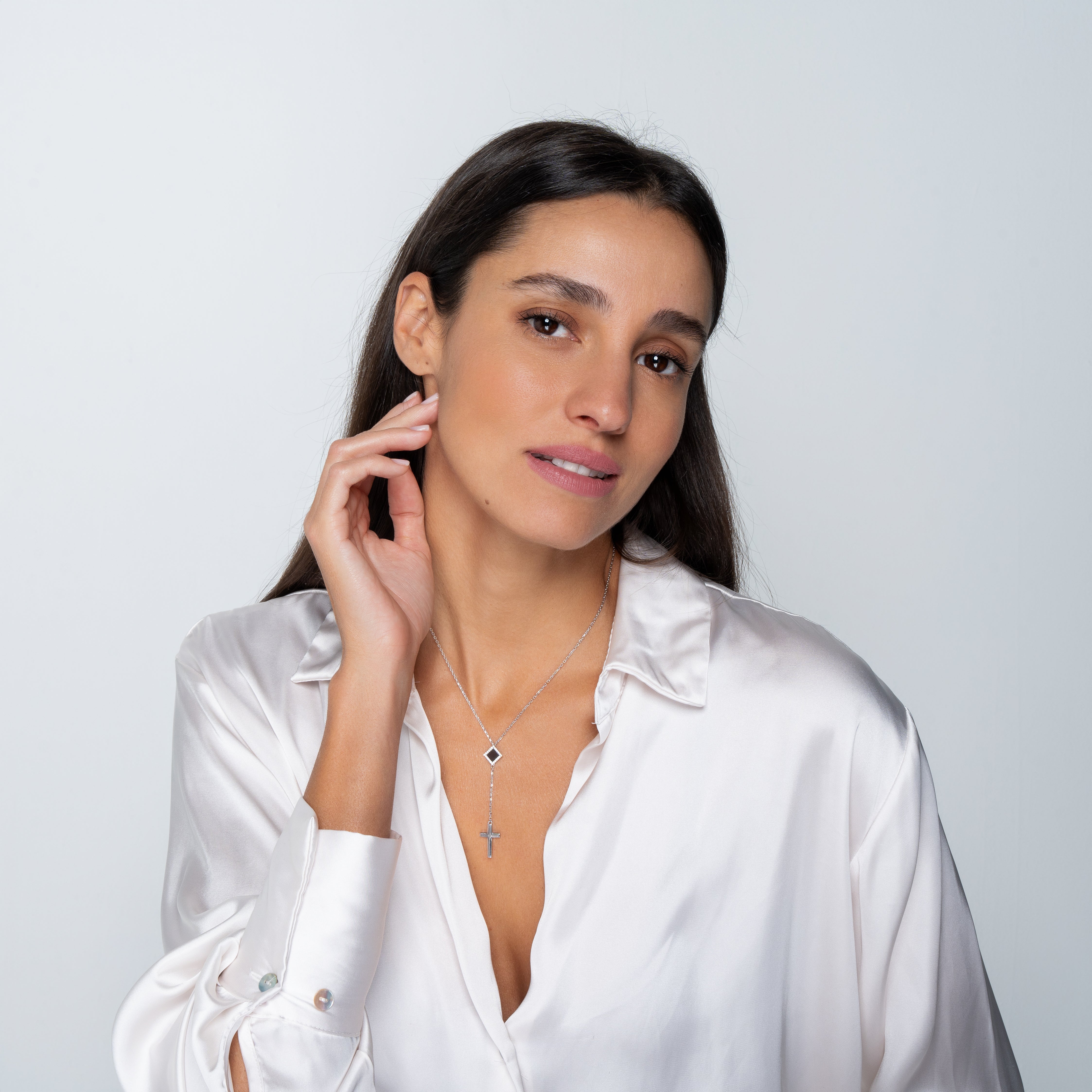 A woman with long dark hair wears a white satin blouse and two silver necklaces, including the Mini Cross Y Necklace by Nano Jewelry, as she poses against a plain light background, gently touching her face and looking slightly to the side.